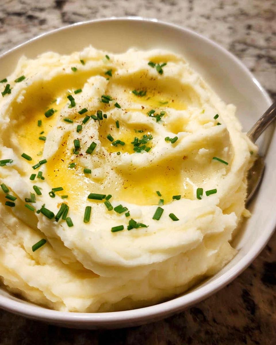 A close-up of fluffy 12-Ingredient Mashed Potatoes topped with melted butter, chives, and pepper.