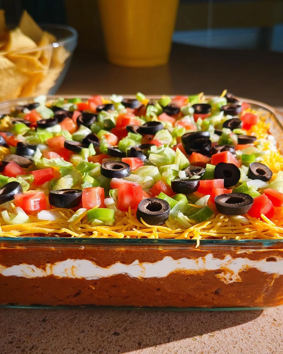 A delicious 7 layer dip with beans, sour cream, cheese, lettuce, tomatoes, and olives, perfect for Taco Tuesday recipes.