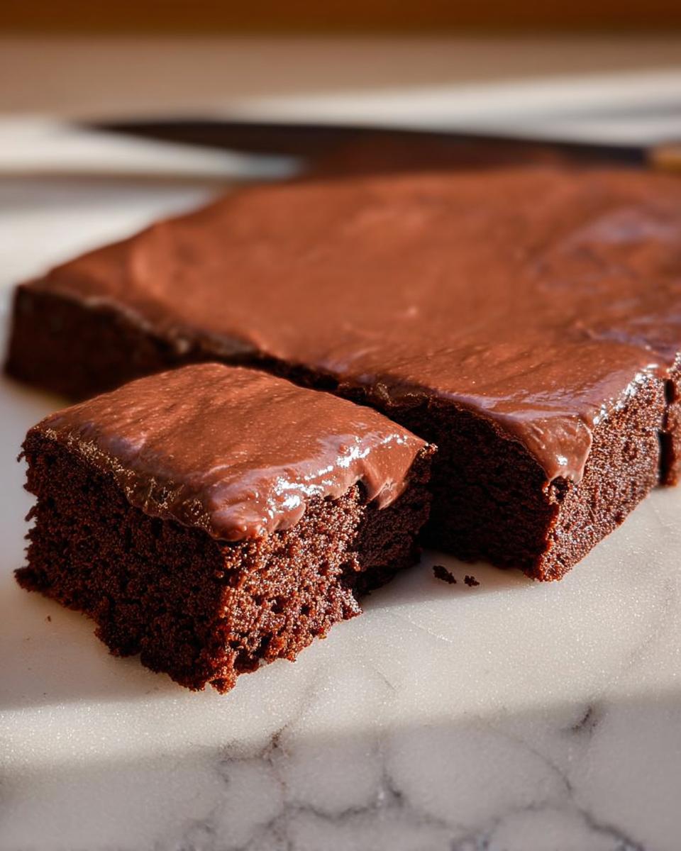 Close-up of a rich chocolate cake, with one slice cut and showing a fluffy texture, frosted with chocolate icing.