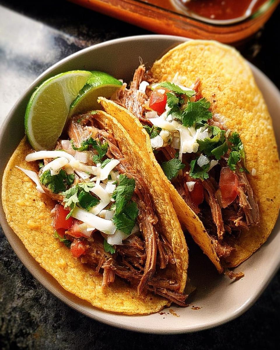 Two corn tortilla tacos filled with shredded beef, topped with diced tomatoes, onions, cheese, cilantro, and a lime wedge.