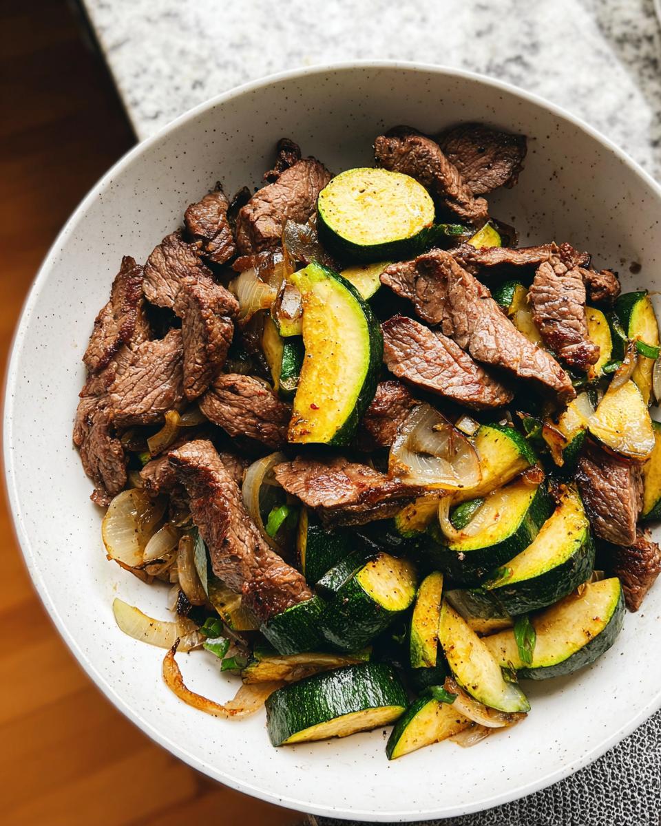 A close-up overhead view of a Cozy Grilled Steak Bowl with Zucchini, featuring sliced steak, zucchini rounds, and onions.