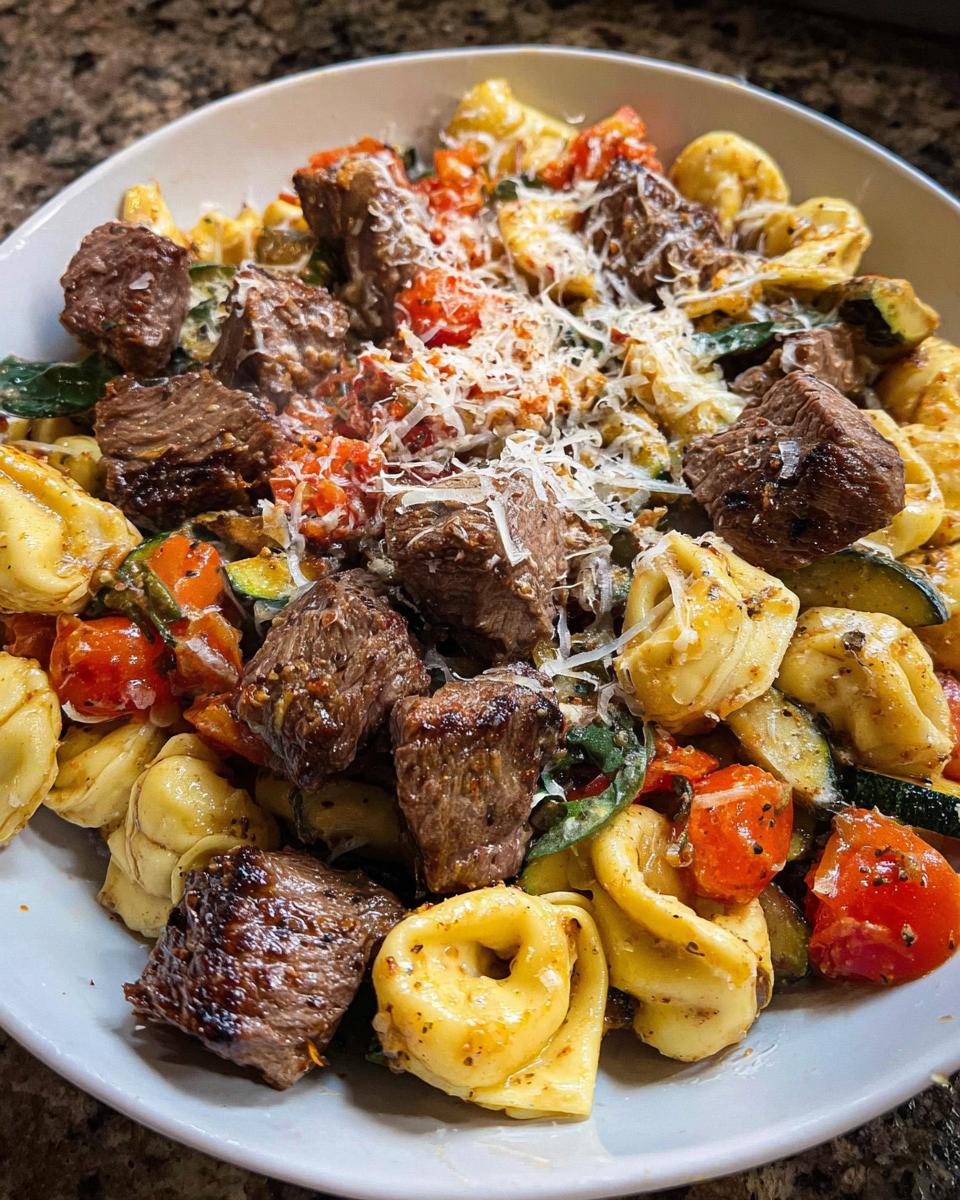 A close-up of a bowl of Garlic Steak Tortellini with chunks of steak, tortellini pasta, tomatoes, and zucchini, topped with shredded cheese.