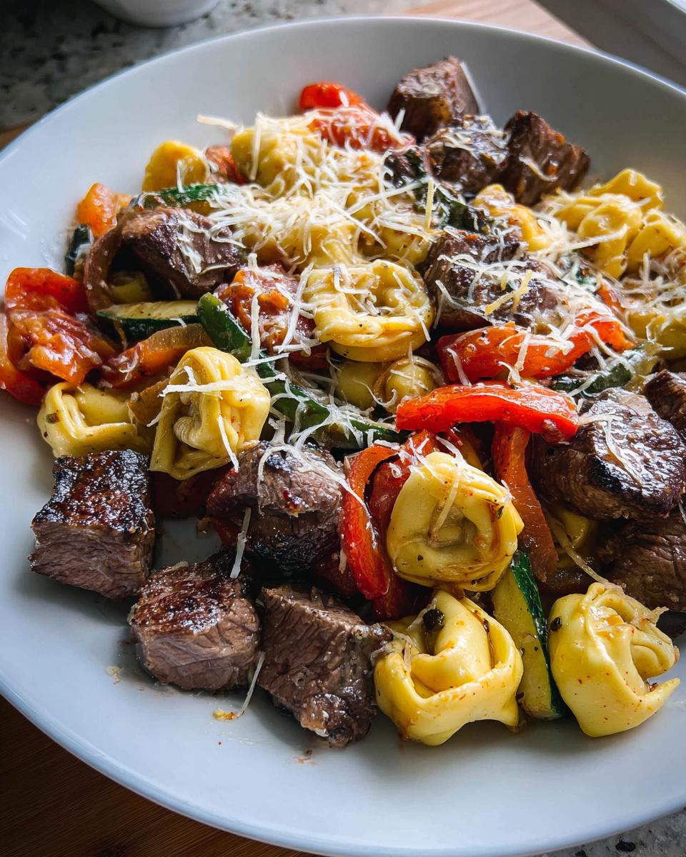 A close-up of a bowl filled with Garlic Steak Tortellini, featuring tender steak bites, cheese tortellini, and colorful bell peppers and zucchini, topped with grated cheese.