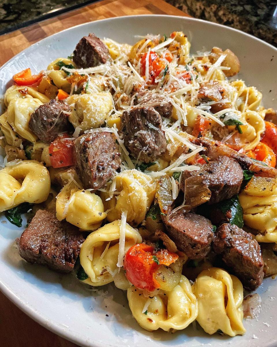 A close-up of a white plate filled with Garlic Steak Tortellini, featuring tender steak pieces, tortellini pasta, cherry tomatoes, and grated Parmesan cheese.