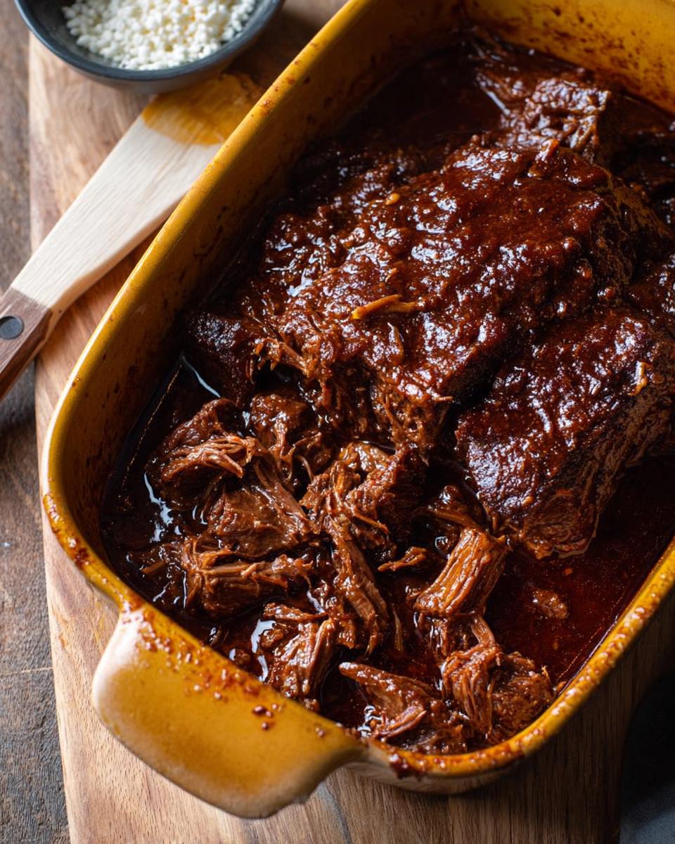 Close-up of a rich, dark sauce covering tender, shredded Korean Style Pot Roast in a yellow baking dish.