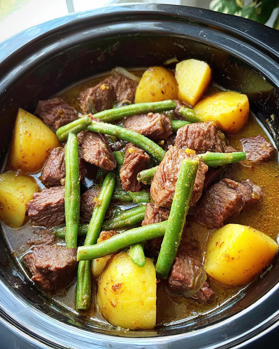 A close-up overhead shot of Slow Cooker Garlic Butter Beef with Potatoes and green beans in a black slow cooker.