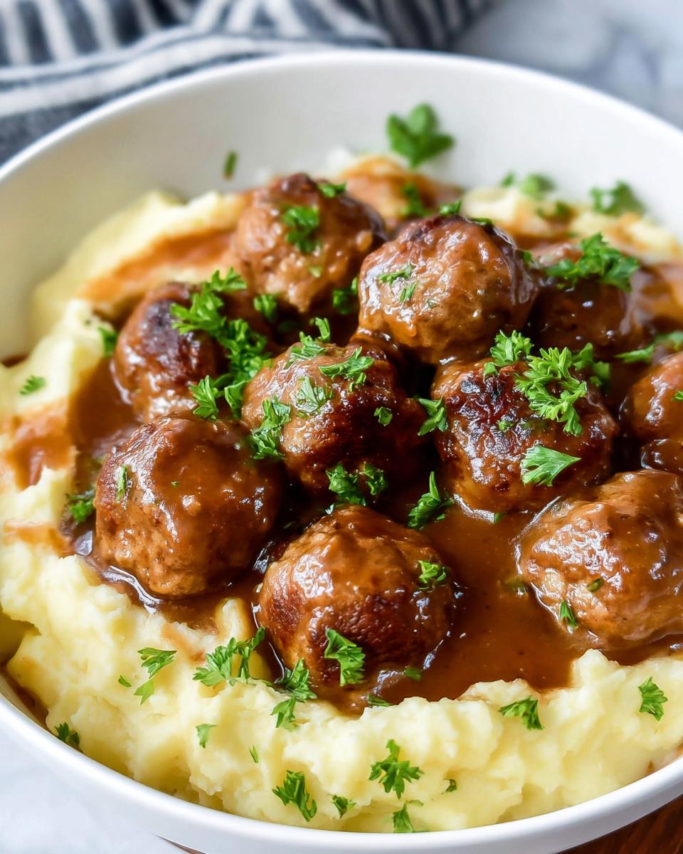 A bowl of Salisbury steak meatballs smothered in brown gravy, served over creamy garlic herb mashed potatoes and garnished with parsley.