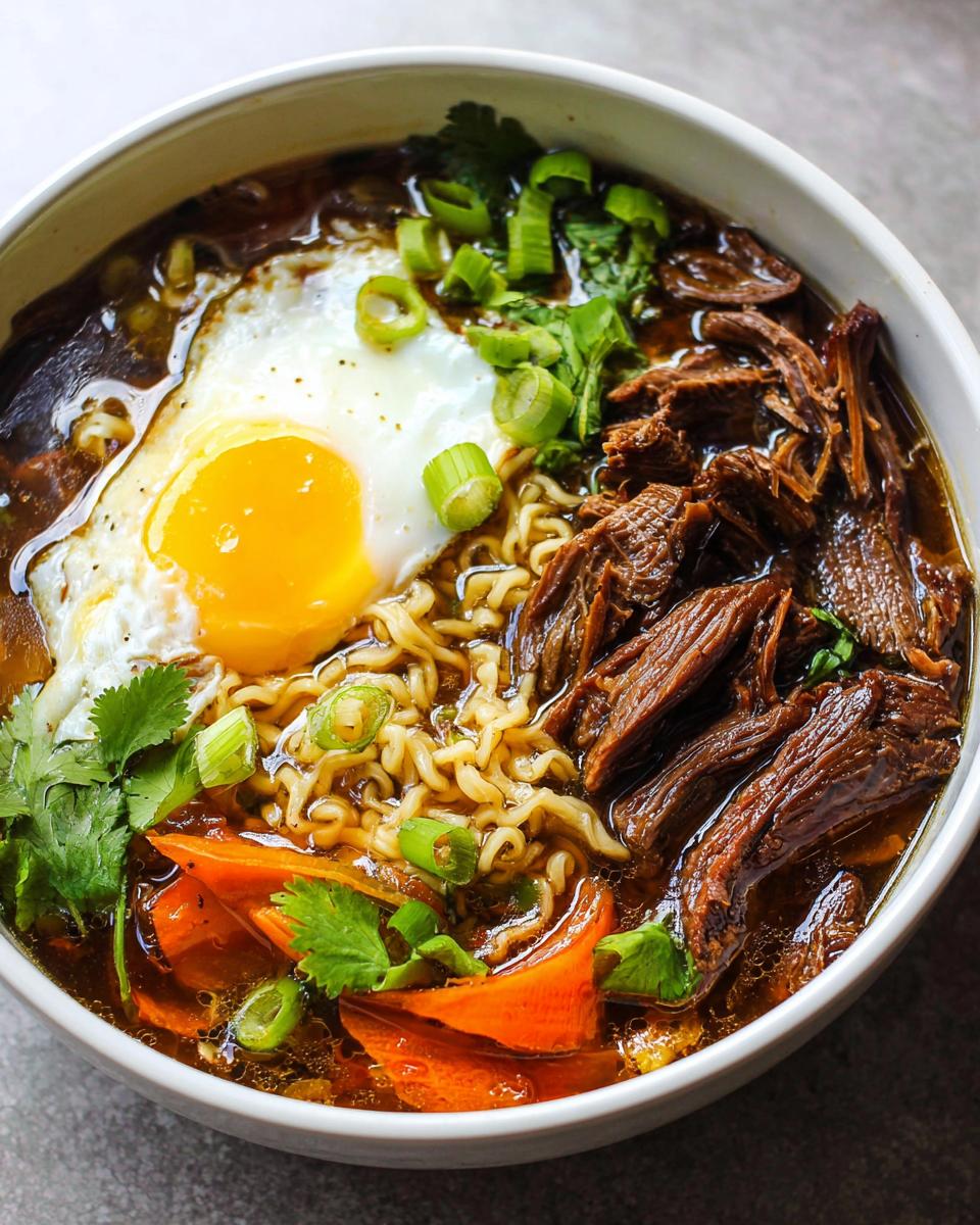 A bowl of Slow Cooker Beef Ramen Noodles topped with a fried egg, shredded beef, carrots, and green onions.