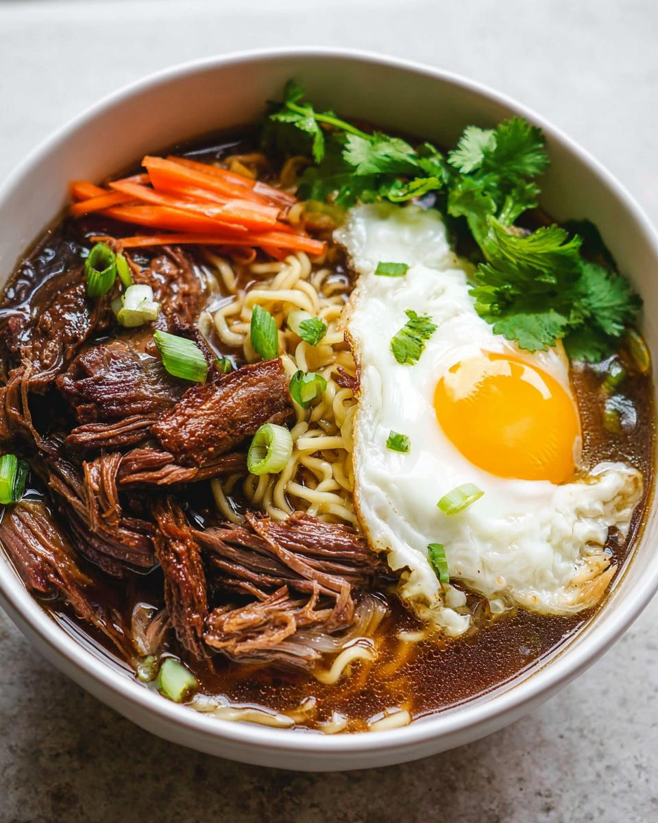 A bowl of Slow Cooker Beef Ramen Noodles topped with a fried egg, shredded beef, carrots, and scallions.