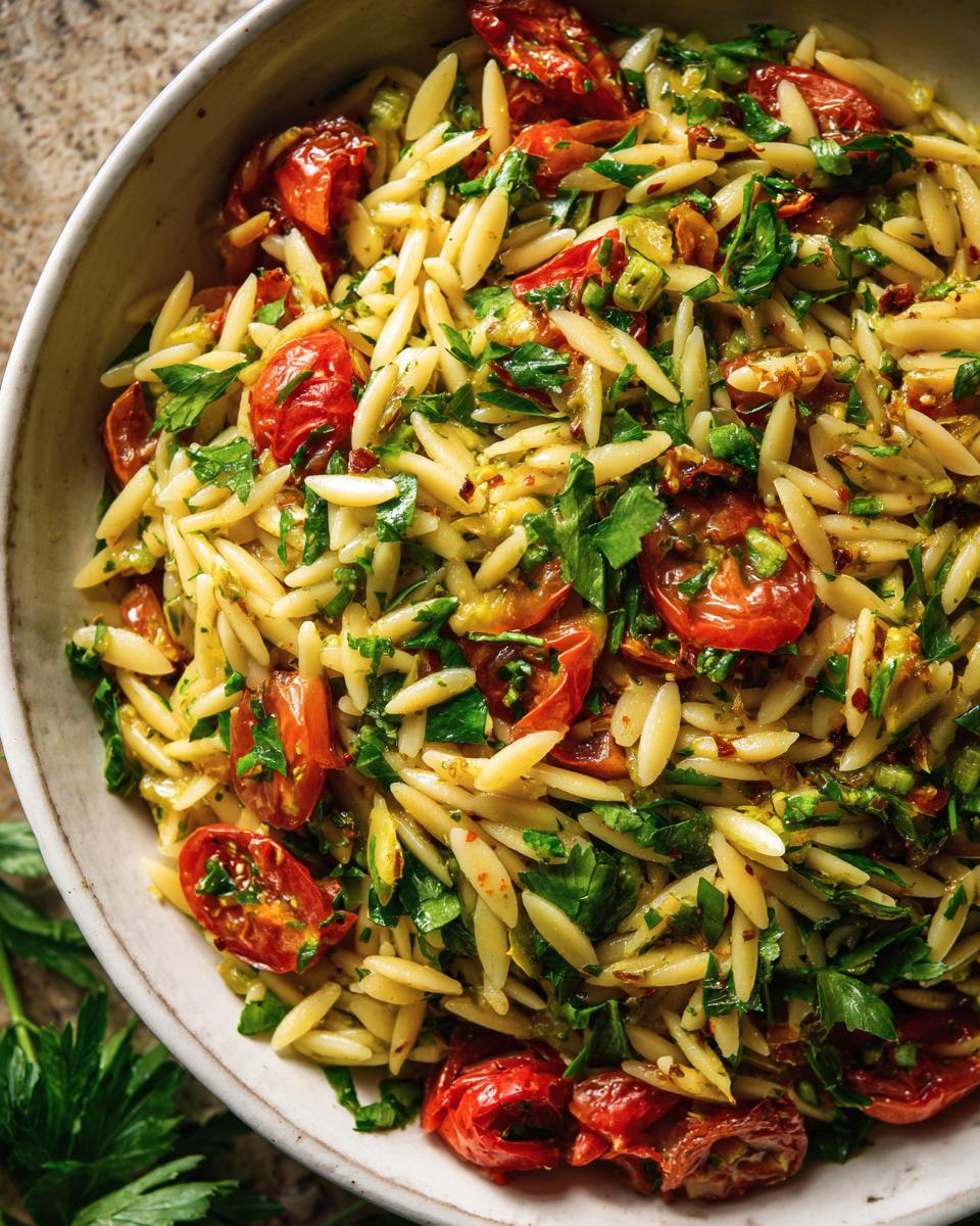 A close-up overhead view of a bowl filled with lemony orzo salad, featuring cherry tomatoes, parsley, and other fresh vegetables.